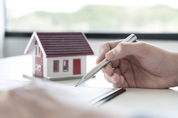 Hand hält einen Stift neben Papierkram auf einem Schreibtisch, im Hintergrund ein kleines Modellhaus. - © Puwasit Inyavileart - stock.adobe.com
