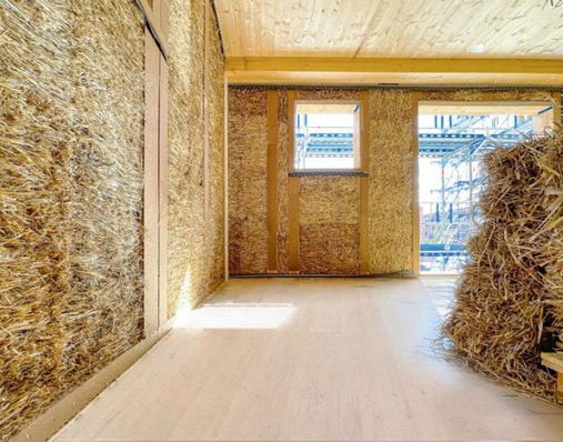 Innenansicht eines im Bau befindlichen Hauses mit Strohballenwänden, Holzbalken und zwei großen Fenstern, außen ein Gerüst. - © Bild: FNR/Dirk Scharmer