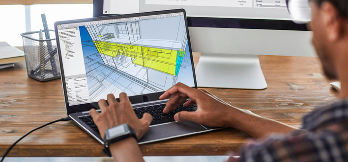 Person, die an einem Laptop an einem Holzschreibtisch mit einem größeren Monitor im Hintergrund an einem architektonischen 3D-Modell arbeitet. - © Bild: Seventyfour - stock.adobe.com