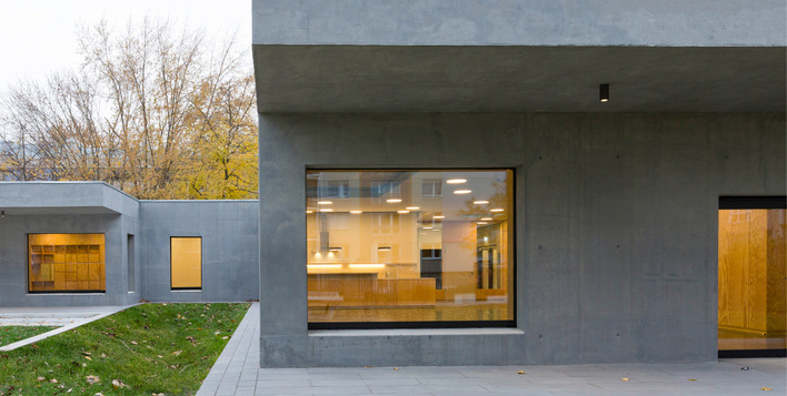 Modernes graues Betongebäude mit großen Fenstern und warmer Innenbeleuchtung, umgeben von Gras und Bäumen. - © Bild: Alexander Blumhoff