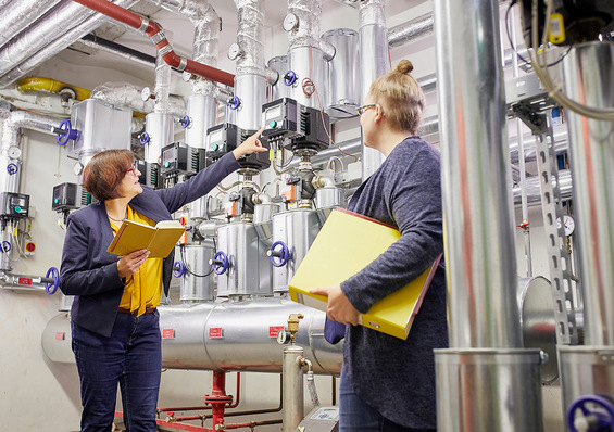 Zwei Personen mit Ordnern prüfen und besprechen Geräte in einem hellen Industrie- oder Maschinenraum. - © Bild: AMX Studio / KEA BW
