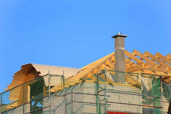 Hölzerner Dachstuhl im Bau mit grünem Sicherheitsnetz und einem Schornstein vor einem klaren blauen Himmel. - © Kara - stock.adobe.com